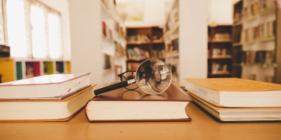 Book in library with old open textbook, stack piles of literature text archive on reading desk, and aisle of bookshelves in school study class room background for academic education learning concept